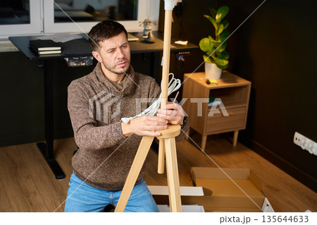 Man assembling floor lamp with electric cable in home interior with unpacked boxes 135644633