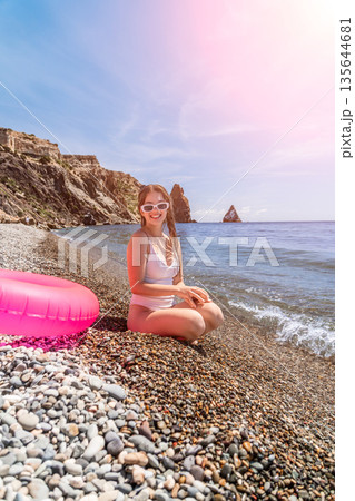 Beach woman vacation young woman in white swimsuit and sunglasses sitting on a pebble beach near the ocean enjoying summer holiday 135644681