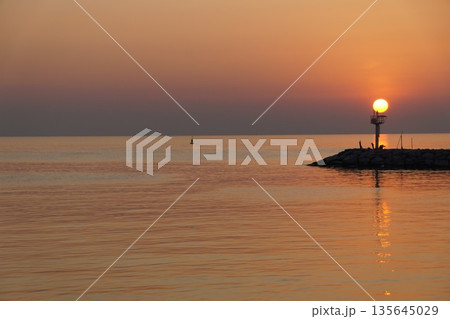 A huge ball of the sun, orange in color, rises from behind the horizon, against the backdrop of a stone pier and a lighthouse.Dawn at sea. 135645029
