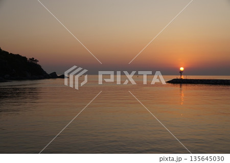 A huge ball of the sun, orange in color, rises from behind the horizon, against the backdrop of a stone pier and a lighthouse. 135645030