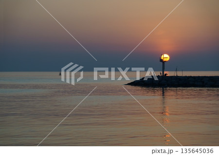 A huge ball of the sun, orange in color, rises from behind the horizon, against the backdrop of a stone pier and a lighthouse. A huge ball of the sun, orange in color, rises from behind the horizon, against the backdrop of a stone pier and a lighthouse. 135645036