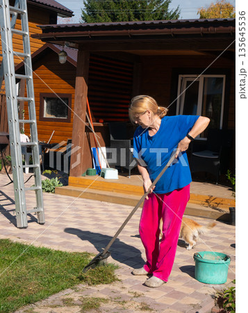 Woman raking cut grass and fallen leaves from a paved patio next to wooden house in a yard during spring or autumn. Woman raking cut grass and fallen leaves from a paved patio next to wooden house in a yard during spring or autumn. 135645536