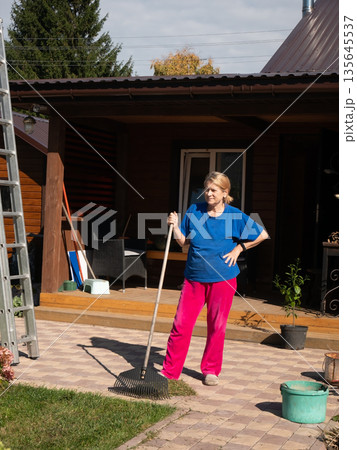 Woman stands with rake on paved patio beside house, ready for autumn garden cleanup work 135645537