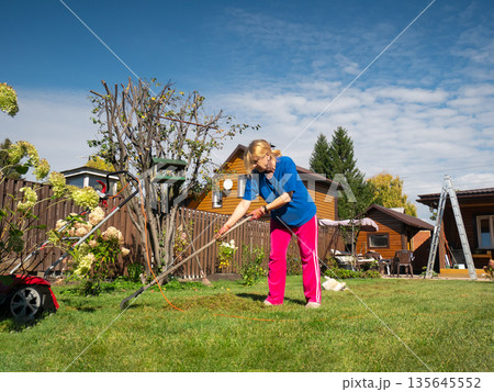 Woman raking cut grass in a lush green backyard garden near a wooden house. Summer outdoor work and home improvement concept. Woman raking cut grass in a lush green backyard garden near a wooden house. Summer outdoor work and home improvement concept. 135645552