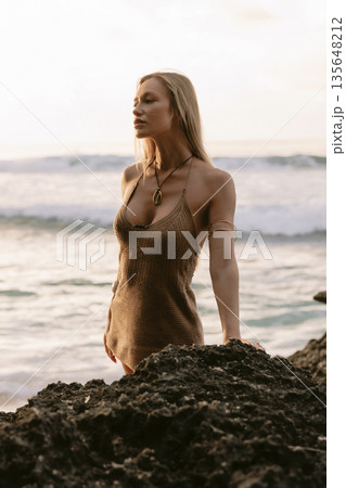Blonde woman in knit dress standing on rocks by the ocean at sunset Blonde woman in knit dress standing on rocks by the ocean at sunset 135648212