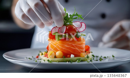 Chef wearing white gloves decorating a gourmet salmon dish with arugula, radish, cherry tomatoes and other fresh ingredients, adding a final touch of elegance to culinary creation 135648367