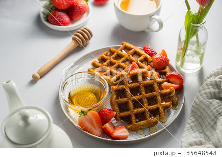 Belgian waffles with strawberries and honey, cup of tea and fresh fruits. Homemade breakfast table setting with tulip flower bouquet Belgian waffles with strawberries and honey, cup of tea and fresh fruits. Homemade breakfast table setting with tulip flower bouquet 135648548