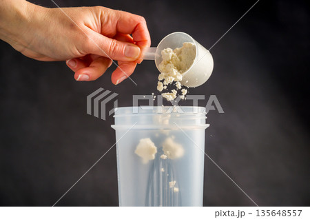 Protein drink. Hand pouring protein powder in a white shaker. Dark background. Body building, muscle growth and food supplement concept 135648557