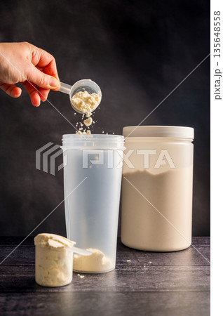 Protein drink. Hand pouring protein powder in a white shaker. Plastic tube on dark background. Body building, muscle growth and food supplement concept 135648558