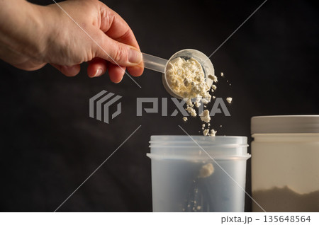 Protein drink. Hand pouring protein powder in a white shaker. Plastic tube on dark background. Body building, muscle growth and food supplement concept 135648564
