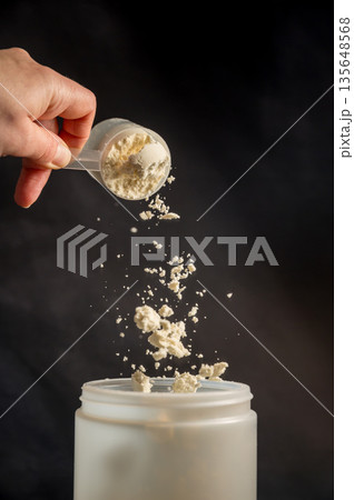 Protein drink. Hand pouring protein powder in a white shaker. Dark background. Body building, muscle growth and food supplement concept Protein drink. Hand pouring protein powder in a white shaker. Dark background. Body building, muscle growth and food supplement concept 135648568