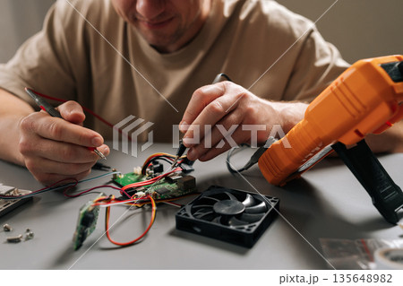 Close-up of technician male repairing electronics, inspecting and testing circuit board with multimeter while surrounded by various hardware components and cooling fan, performing expert maintenance. Close-up of technician male repairing electronics, inspecting and testing circuit board with multimeter while surrounded by various hardware components and cooling fan, performing expert maintenance. 135648982