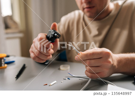 Man heats stripped electrical cable with gas torch while soldering and repairing electronics at cluttered workbench, focused on precise diy wiring and connection work. Man heats stripped electrical cable with gas torch while soldering and repairing electronics at cluttered workbench, focused on precise diy wiring and connection work. 135648997