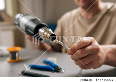 Close-up of person hands working on damaged electronic cable, applying heat with heat gun to shrink tubing or solder connecting wires, signifying diligent repair work and technical expertise. Close-up of person hands working on damaged electronic cable, applying heat with heat gun to shrink tubing or solder connecting wires, signifying diligent repair work and technical expertise. 135649003
