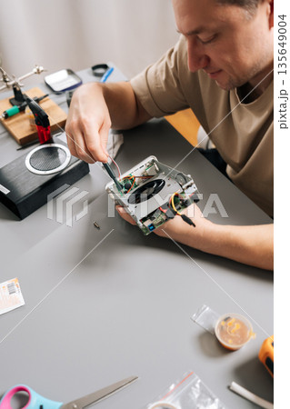 Top view of technician male working on electronic device, holding computer hardware with exposed cooling fan, performing repairs and maintenance on tech equipment in modern workshop. Top view of technician male working on electronic device, holding computer hardware with exposed cooling fan, performing repairs and maintenance on tech equipment in modern workshop. 135649004