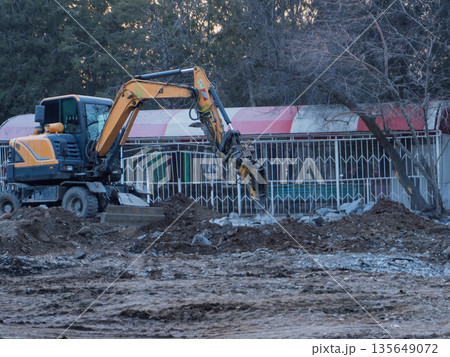 Yellow excavator with hydraulic breaker working on a construction site. Heavy machinery demolishing ground with jackhammer attachment 135649072