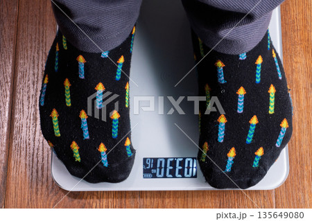 Top view of feet in socks standing on a digital weighing scale. Person measuring body weight of 103 kg on wooden floor. Obesity and diet concept 135649080