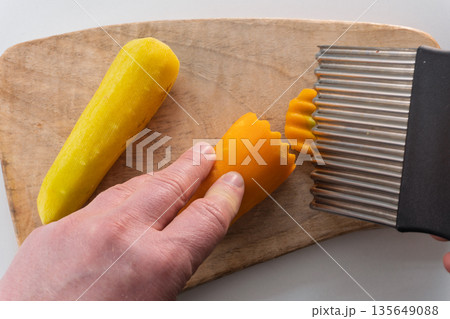 Person slicing raw yellow carrot with crinkle cutter on wooden cutting board. Top view of hand preparing fresh vegetables with wavy knife. Healthy cooking concept 135649088