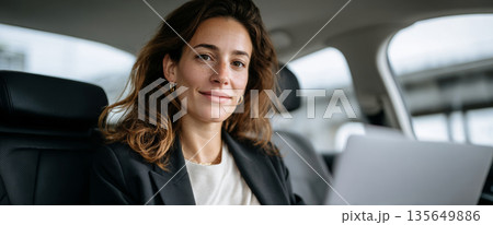 Confident young woman sitting in car backseat working on laptop with relaxed expression and casual business attire 135649886