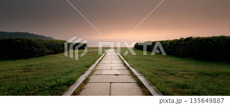 Symmetrical stone pathway leading through green grass and trimmed hedges towards a calm horizon under a soft orange sky at dusk 135649887