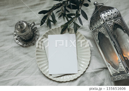 Ramadan Blank Greeting Card on Ceramic Plate, Fabric Table Background. Silver Lantern, Arabic Coffee Cup. Olive Branch on Fabric Background. Festive Islamic Mockup for Eid Invitations. Flat lay, Top Ramadan Blank Greeting Card on Ceramic Plate, Fabric Table Background. Silver Lantern, Arabic Coffee Cup. Olive Branch on Fabric Background. Festive Islamic Mockup for Eid Invitations. Flat lay, Top 135651606