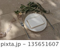 Elegant wedding setting flat lay. Blank place card on ceramic plate, olive branch decor, beige marble floor. Rustic Mediterranean summer neutral dining scene. Minimalist stationery mockup copy space. 135651607