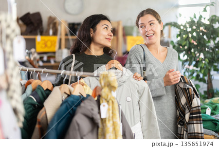 Two happy young women choosing coat or jacket for cold season in clothing shop with large assortment Two happy young women choosing coat or jacket for cold season in clothing shop with large assortment 135651974