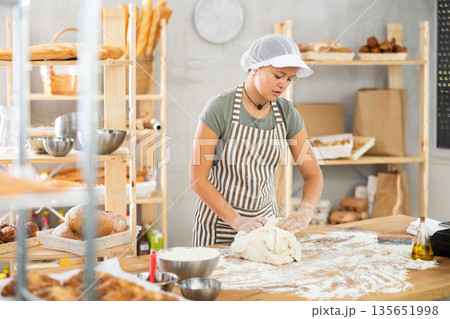 Young smiling female baker preparing raw yeast dough on kitchen table in bakery Young smiling female baker preparing raw yeast dough on kitchen table in bakery 135651998