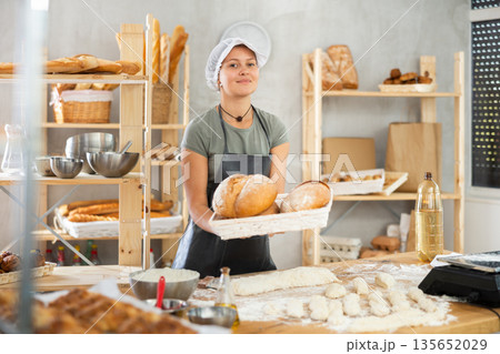 Young woman baker are standing near product range in bakery kitchen with basket of bread. Young woman baker are standing near product range in bakery kitchen with basket of bread. 135652029