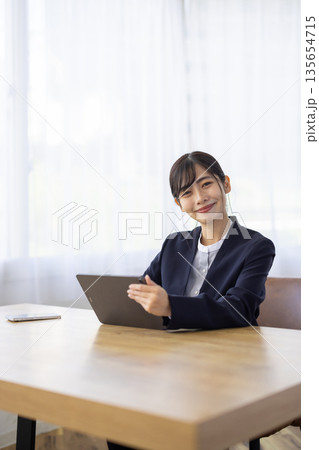 タブレットを操作しながらカメラ目線で微笑むスーツを着た若い女性 135654715