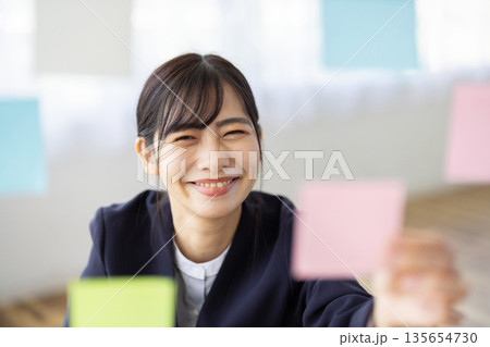 オフィスで付箋を手に微笑むスーツを着た若い女性 オフィスで付箋を手に微笑むスーツを着た若い女性 135654730