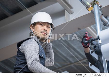 天井が近い高所の足場で単管パイプを固定するクランプなどの施工作業　鳶職や作業員のイメージカメラ目線 135656335