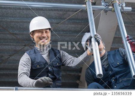 天井が近い高所の足場で単管パイプを固定するクランプなどの施工作業　鳶職や作業員のイメージ笑顔 135656339