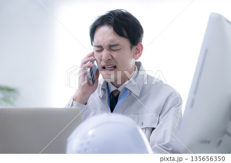 現場事務所で電話対応する作業員　発注や下請けで納期に困る　スマホでクレームの応対	 135656350