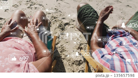 Sitting senior couple viewing floating social media icons on wooden deck chairs on sandy beach 135657598