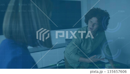 Gesturing man with curly hair leaning forward at meeting room table, with notebooks and monitor 135657606
