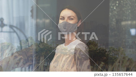 Standing woman wearing grey cloth mask and white top at window, reflecting grass and skyline Standing woman wearing grey cloth mask and white top at window, reflecting grass and skyline 135657644