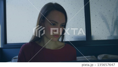 Working woman wearing burgundy top typing on laptop on sofa in living room, with rain-soaked window Working woman wearing burgundy top typing on laptop on sofa in living room, with rain-soaked window 135657647