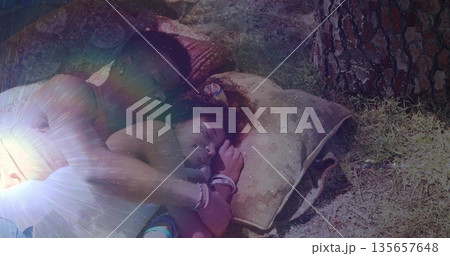 Lying couple cuddling on pillows on grass by tree, wearing tank and sleeveless tops and bracelets 135657648