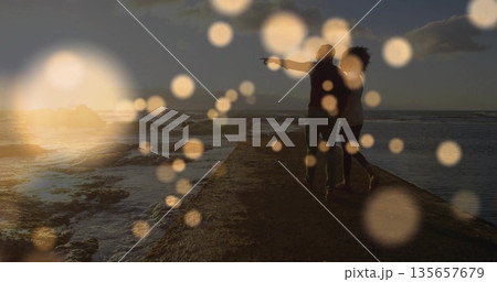 Walking couple pointing toward ocean rocks at sunset with bokeh orbs on stone pier, copy space 135657679