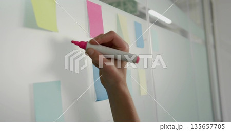 Holding hand gripping pink marker poised on glass board in office with pastel notes, copy space 135657705
