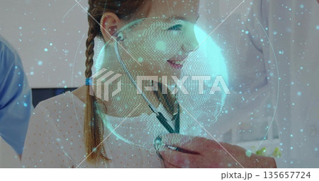 Lab coat doctor placing stethoscope on girl in clinic, with blue scrubs staff and hologram globe 135657724