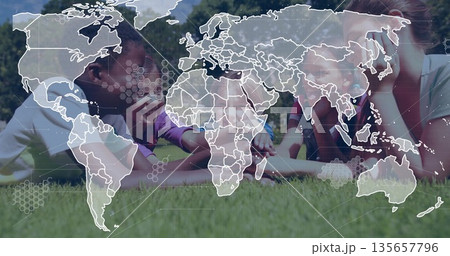 Lying diverse children studying tablet computer on lush green lawn in park, with world map overlay 135657796