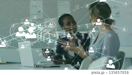Two businesswomen in blazers collaborating using laptops and tablet in office, with network overlay 135657858