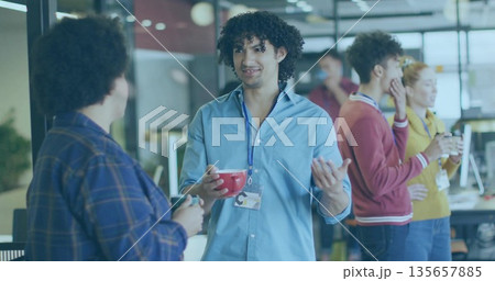Gesturing man in light blue shirt ID badge holding red mug talking in break area 135657885