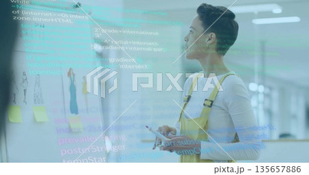 Examining whiteboard sketches and code, woman in overalls holding stylus and phone at design studio 135657886