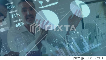 Analyzing business team in suits gesturing toward data overlay in office, with laptops and phone 135657931
