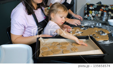 Young mother and her daughters cut ginger cookie dough and place the shaped pieces on a baking tray 135658574