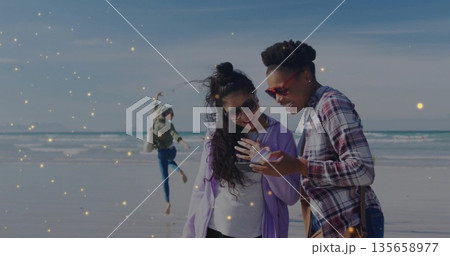 Leaping woman and friends leaning sharing smartphone on beach shoreline, with bags and sunglasses Leaping woman and friends leaning sharing smartphone on beach shoreline, with bags and sunglasses 135658977
