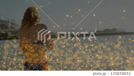 Standing woman wearing blue bikini at sea edge by breakwater, with bokeh light reflections Standing woman wearing blue bikini at sea edge by breakwater, with bokeh light reflections 135659032